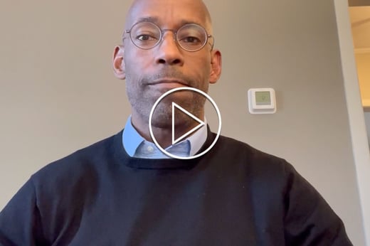 A portrait of Michael Collins wearing a dark sweater and blue dress shirt. There is a white stenciled play button in the fore front. 