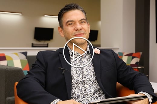 A protrait of Wil Del Pilar, Ph.D. wearing a dark blazer and black and white dress shirt. There is a white stenciled play button in the fore front.