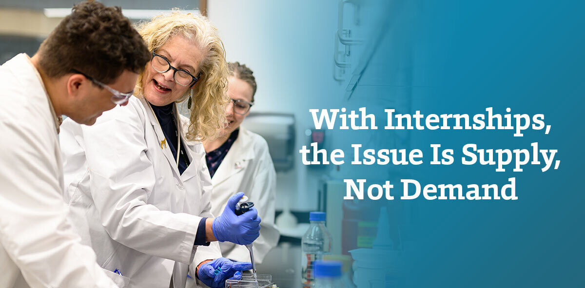In white, text says with internships, the issue is supply, not demand. To the right is a professor and their students, in white lab coats, and safety goggles interacting with each other. 